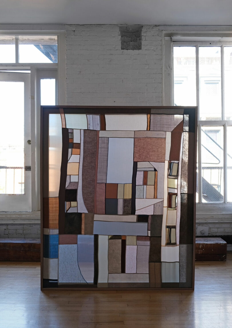 A large abstract textile artwork by Kiva Motnyk with geometric shapes in neutral tones is displayed in front of two tall windows in a bright, industrial-style room.