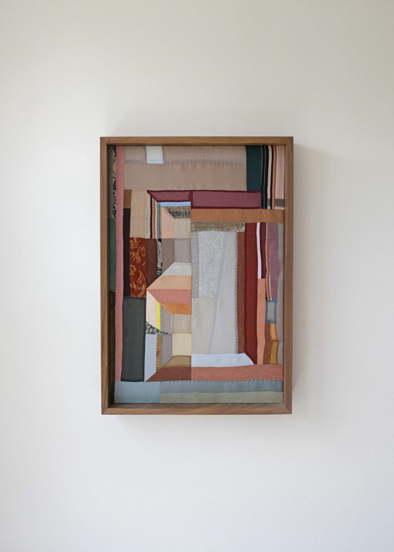 A framed textile artwork by Kiva Motnyk, featuring an abstract geometric pattern crafted from various fabric pieces in muted earth tones, is mounted on a plain white wall.