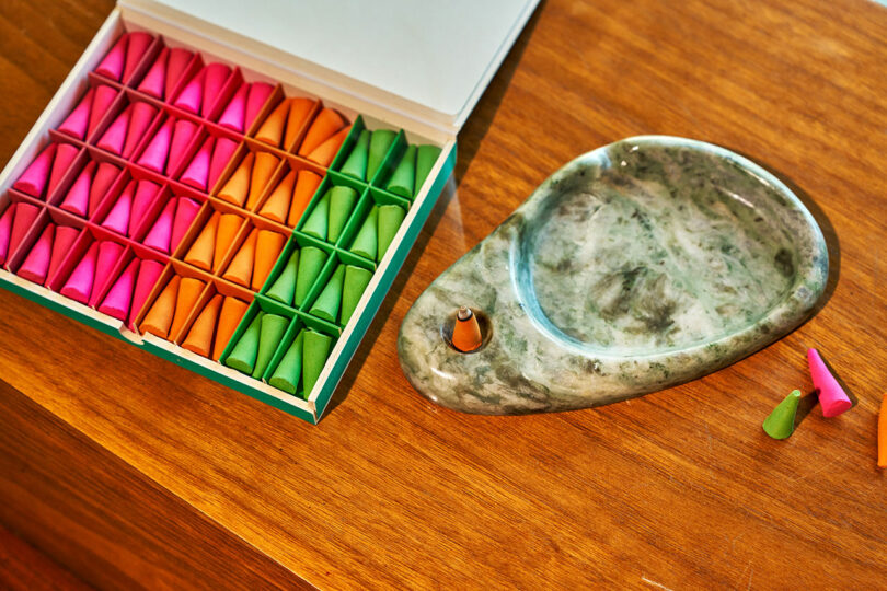 A box of colorful incense cones sits next to a green marble incense holder on a wooden surface, with a few incense cones nearby.