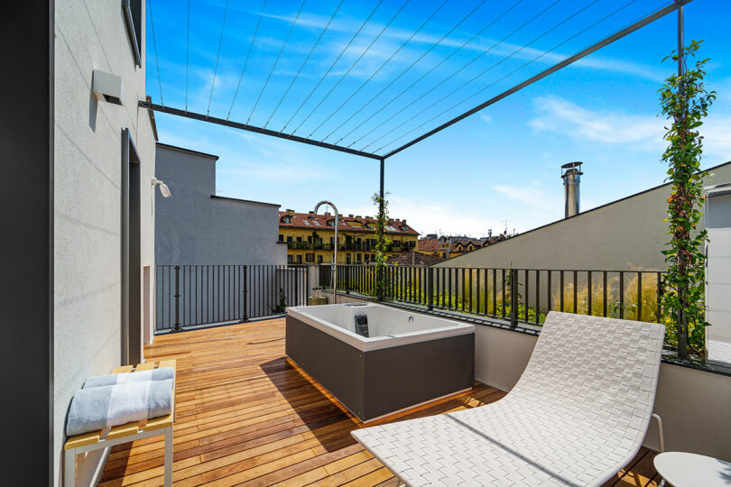 Modern rooftop terrace with wooden decking, a white lounge chair, a square hot tub, towel rack, and metal railing, overlooking neighboring buildings under a blue sky.