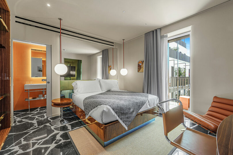 Modern hotel room with a large bed, glass desk, brown leather chair, marble floor, and a view through tall windows; bathroom visible in the background.