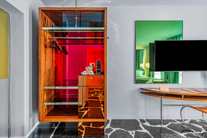 Open wardrobe with glass shelves, hanging space, cups, and a coffee machine next to a desk and wall-mounted TV in a modern hotel room.