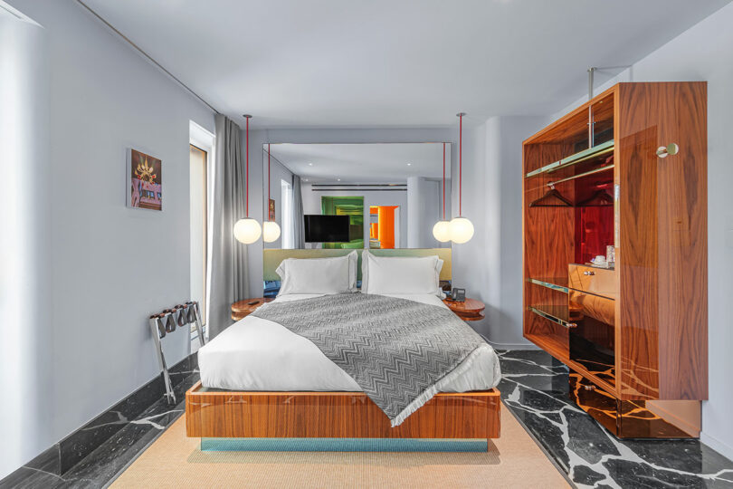 Modern hotel room with a wooden bed, gray blanket, marble floor, glass wardrobe, bedside pendant lights, and a large wall mirror behind the bed.
