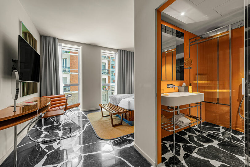 Modern hotel room with black-and-white marble floor, a bed, wooden furniture, large windows, and an orange-accented bathroom with a glass shower enclosure.