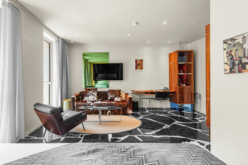 Modern living area with a cowhide sofa, black lounge chair, glass coffee table, wall-mounted TV, wooden desk, marble floor, and large windows with sheer curtains.