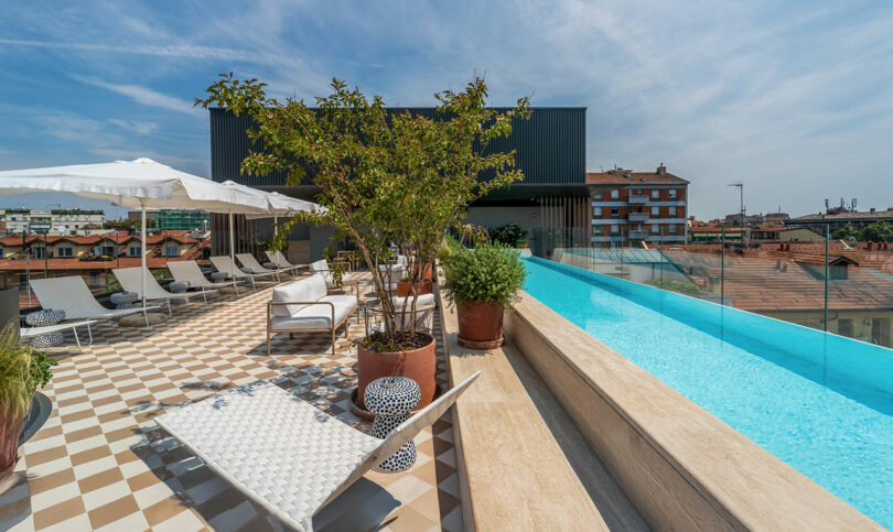Rooftop pool area with lounge chairs, potted plants, and umbrellas overlooking city buildings under a partly cloudy sky.