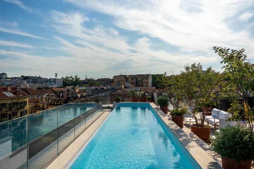 A rooftop swimming pool with glass railing, potted plants, lounge chairs, and a cityscape view under a partly cloudy sky.