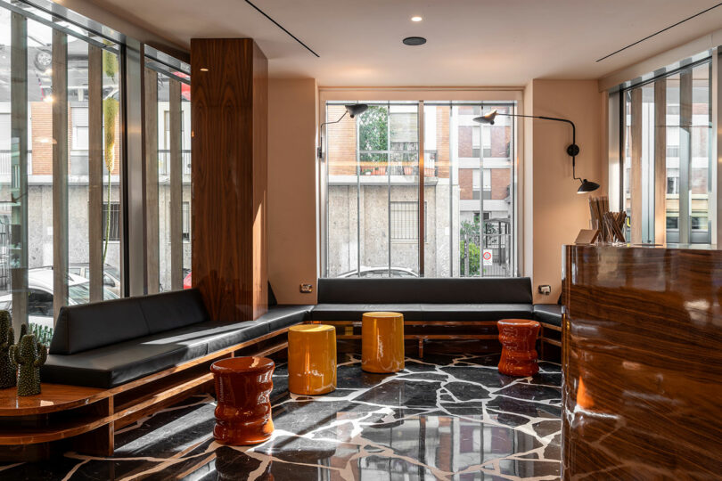 Modern waiting area with black bench seating, amber and red stools, marble-patterned floor, large windows, and wood-paneled surfaces.