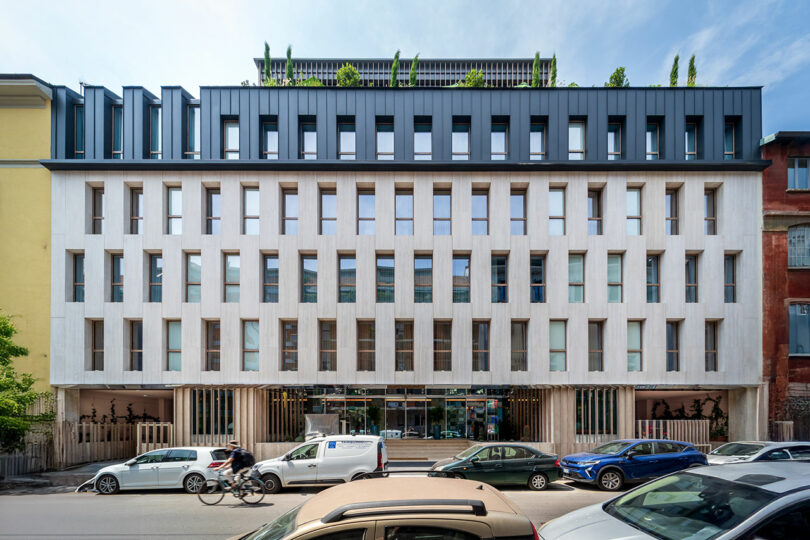 Modern multi-story building with a grid-like facade, large windows, rooftop greenery, and several cars and a cyclist on the street in front.
