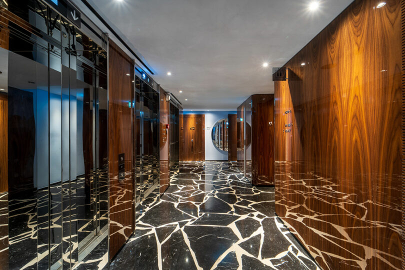 A modern elevator lobby with glossy wood-paneled walls, mirrored surfaces, and a black marble floor with white veining, illuminated by recessed ceiling lights.