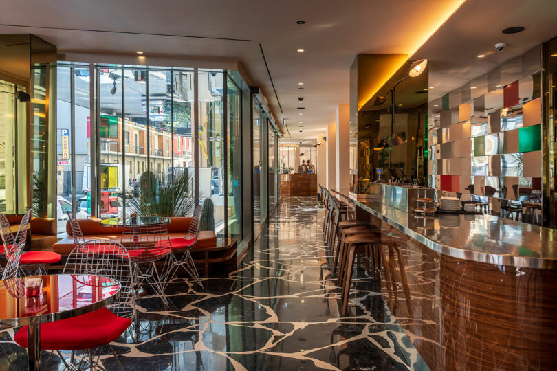 Modern restaurant interior with marble floor, mirrored walls, red tables, wire chairs, and a long wooden bar; large windows show a city street outside.