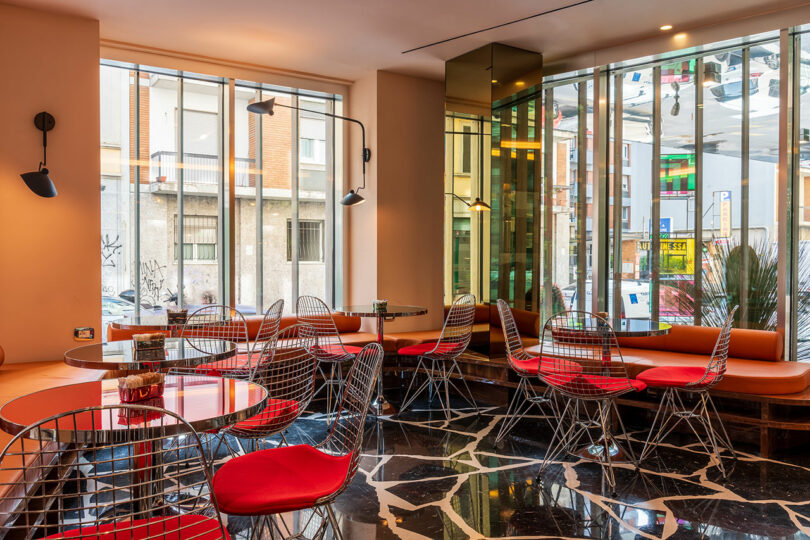 Modern cafe interior with wire chairs featuring red cushions, round tables, large windows, and a black patterned floor. Natural light illuminates the space.