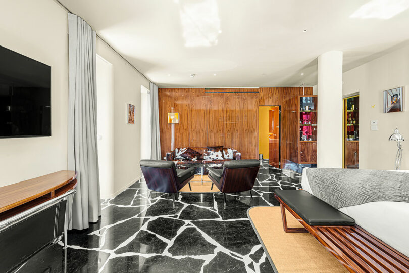 Modern living room with black and white marble floor, wood-paneled wall, black lounge chairs, a glass coffee table, and a mounted TV. Bright, minimalist decor with large windows and shelving.