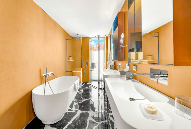 Modern bathroom with orange walls, a freestanding white bathtub, double sinks, mirrored cabinets, glass shower, and black and white marble flooring.