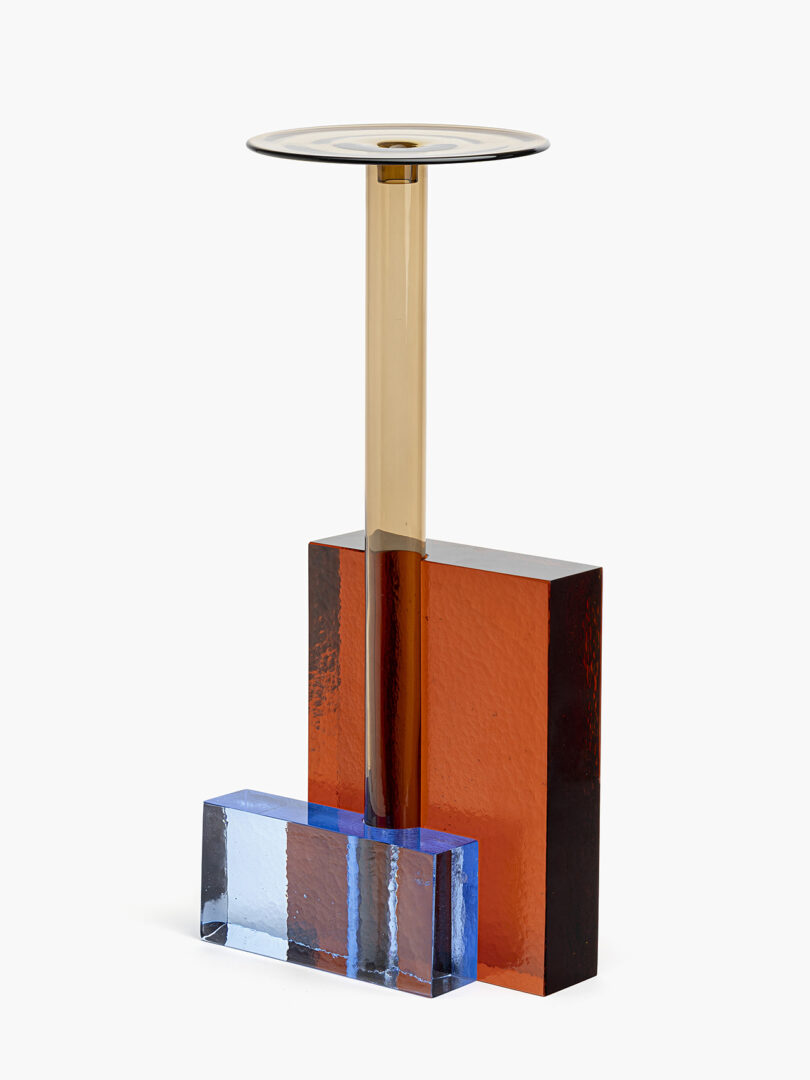 La escultura moderna presenta un soporte cilíndrico dorado, una base rectangular roja y una decoración rectangular azul, todos hechos de vidrio o materiales acrílicos.