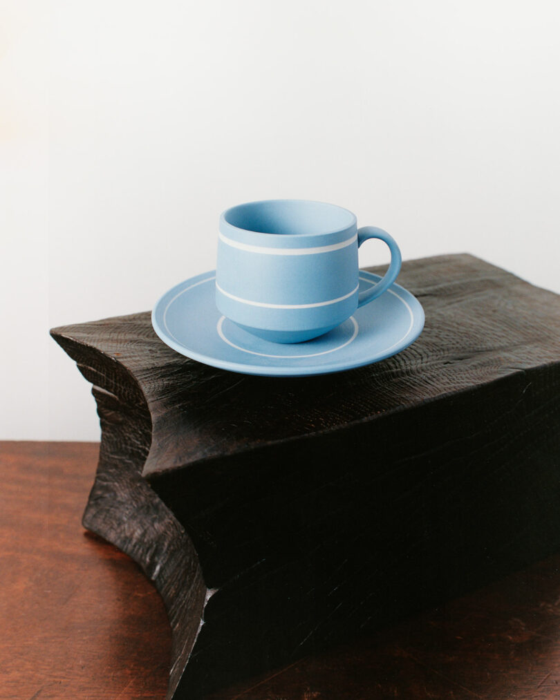 A light blue cup and saucer set is placed on a carved, dark wooden block atop a brown wooden surface, against a plain white background.