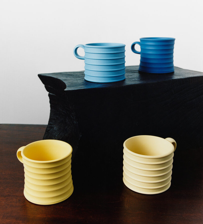 Four ribbed ceramic mugs, two yellow and two shades of blue, are arranged on and in front of a dark, textured block against a plain background.