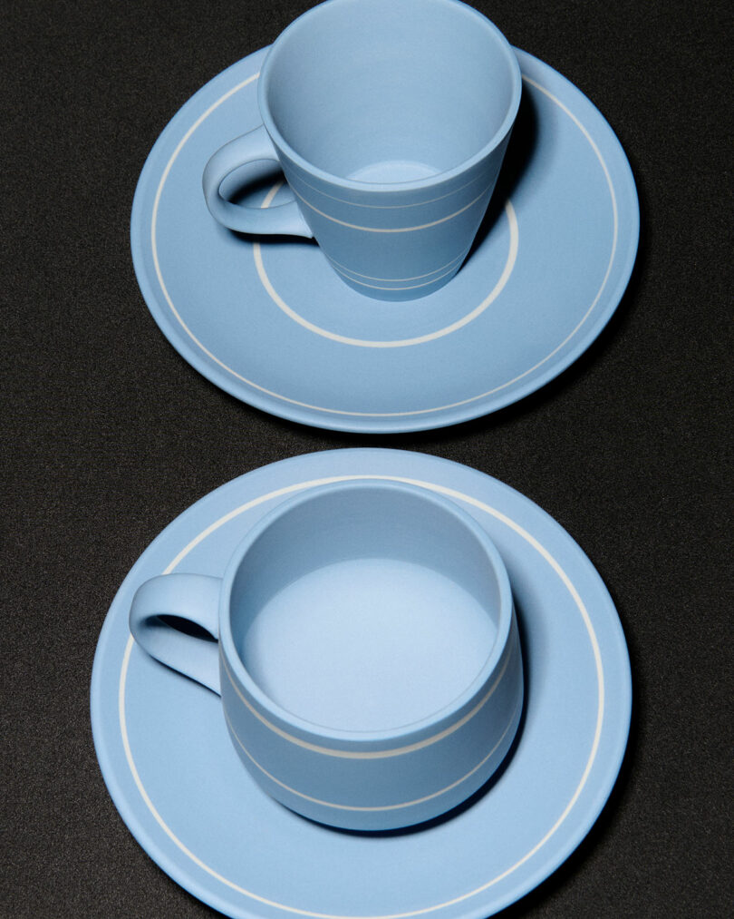 Two light blue ceramic teacups on matching saucers with white circular lines, reminiscent of JW Anderson Wedgwood designs, placed on a dark, textured surface.