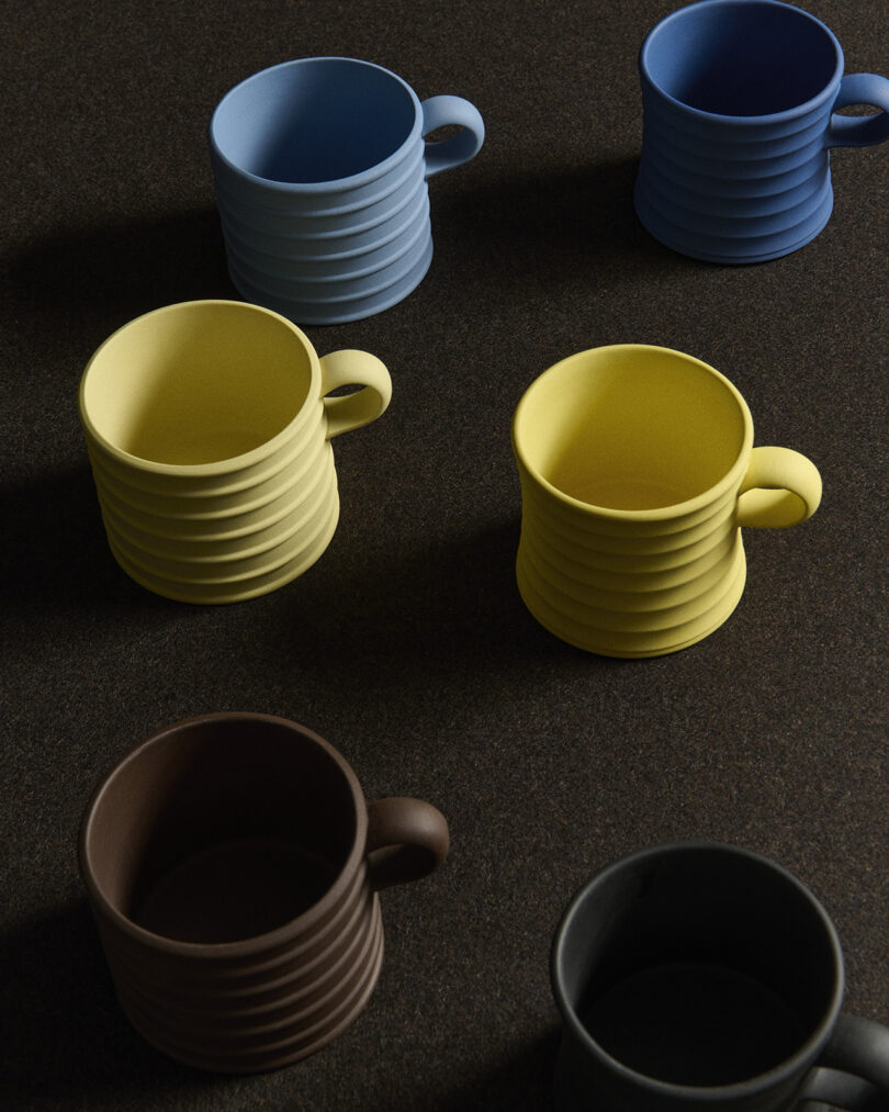 Six ribbed ceramic mugs in yellow, blue, brown, and black are arranged on a dark textured surface, viewed from above.