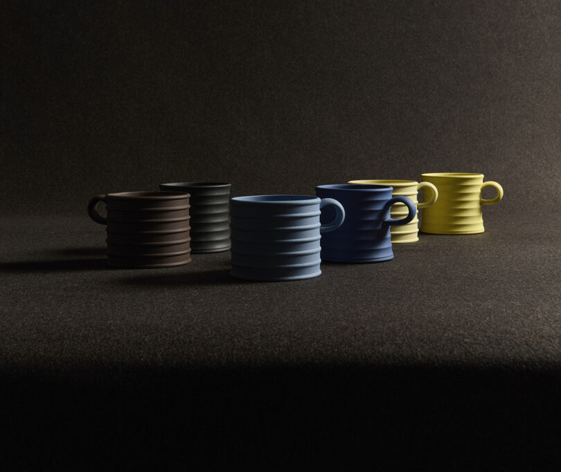 Five ribbed ceramic mugs in brown, black, blue, yellow, and light yellow are arranged in a line on a dark textured surface, with soft lighting from the right.