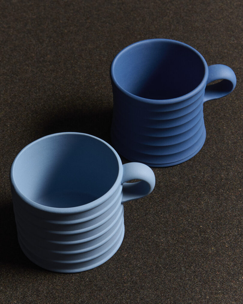 Two ribbed ceramic mugs, one light blue and one dark blue, are placed on a dark textured surface.