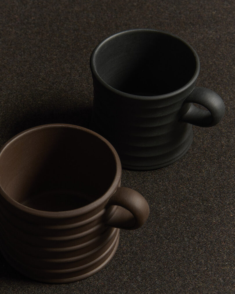 Two ribbed ceramic mugs, one black and one brown, are placed on a dark textured surface.