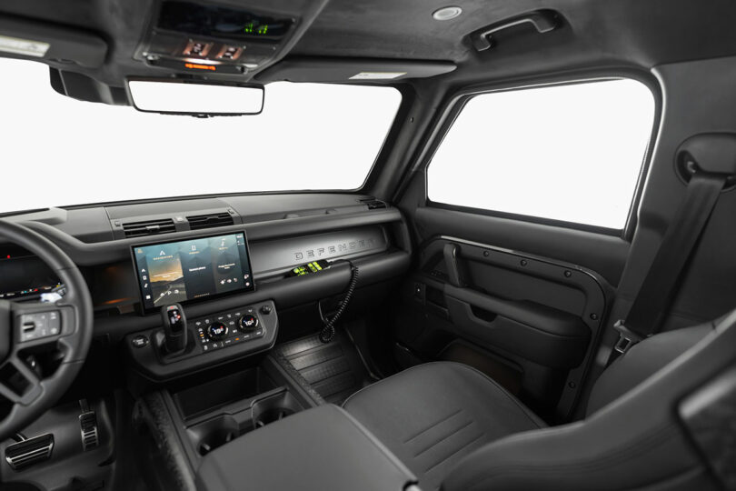 The image shows the interior of a modern vehicle with black seats, a large central touchscreen, joystick controls, and minimalistic dashboard design.