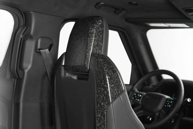 Close-up view of a car interior showing the driver's seat backrest with a speckled pattern, part of the steering wheel, and dashboard controls.