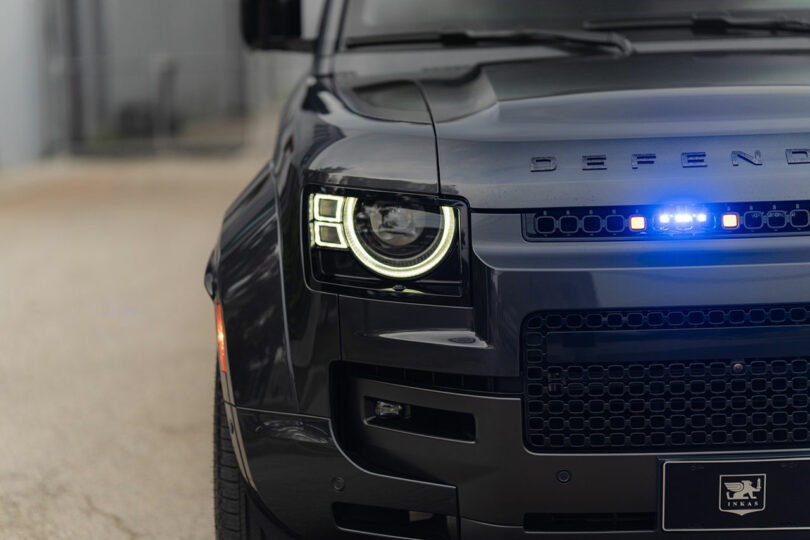 Primer plano del lado delantero izquierdo de un Land Rover Defender negro con faros LED encendidos y una luz azul en la parrilla.