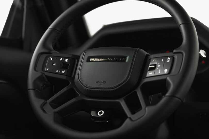 Close-up of a modern black car steering wheel with control buttons and a “Defender” badge in the center.