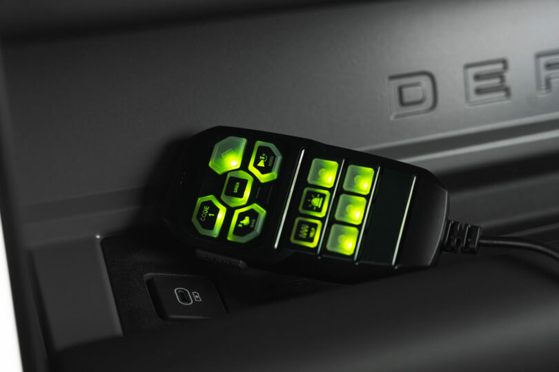 A black electronic control device with illuminated green buttons rests on a textured surface next to an electrical outlet.