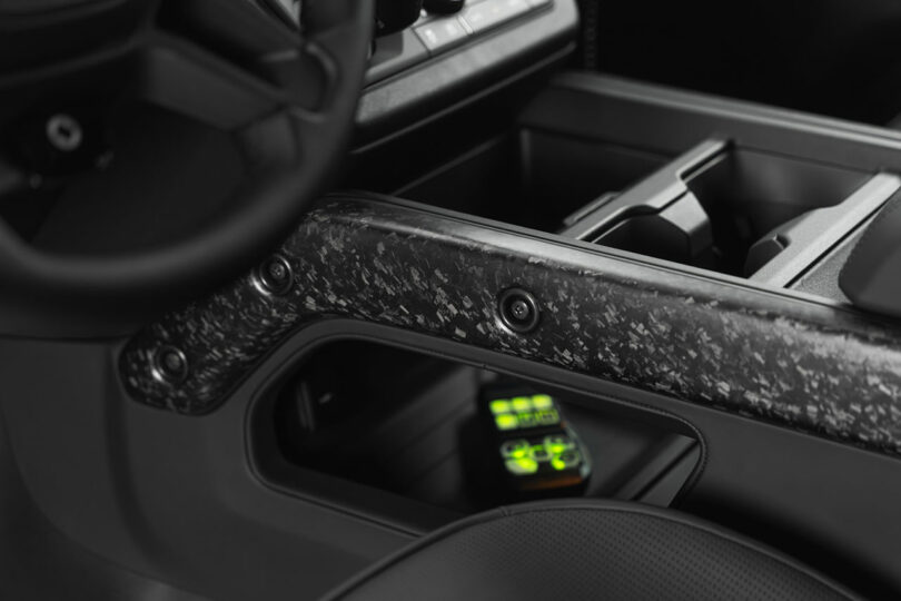 Close-up of a car’s center console with a textured black panel and a small electronic device displaying green lights stored in a compartment.