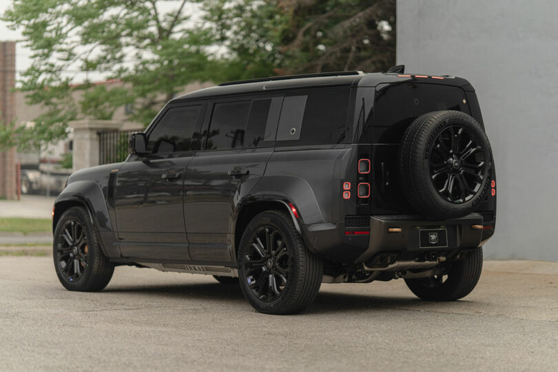 A black SUV with tinted windows and a spare tire mounted on the rear is parked on a street next to a gray wall.