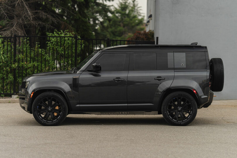 A dark gray SUV with black wheels is parked on a paved surface in front of a fence and greenery.
