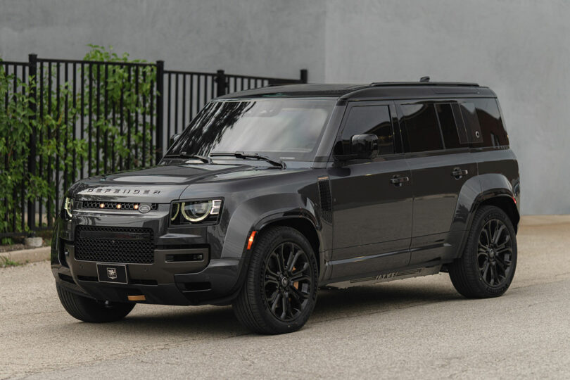 A black Land Rover Defender SUV is parked on a street in front of a metal fence and a gray wall.