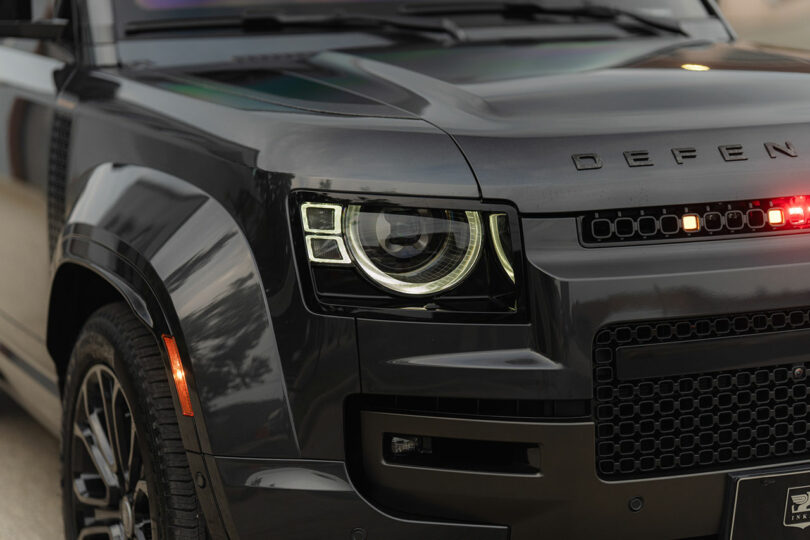 Close-up view of the front left headlight and grille of a black Land Rover Defender, with "DEFEN" lettering visible on the hood.