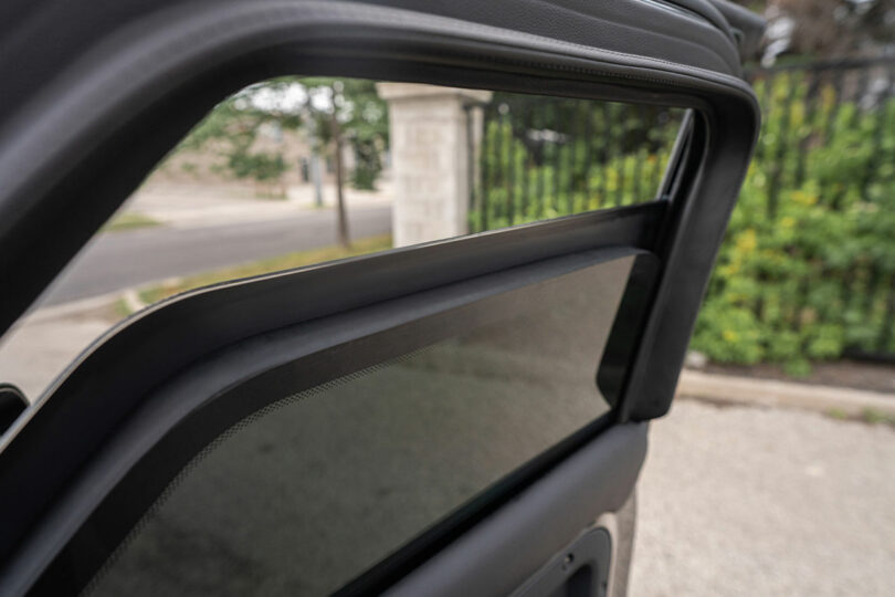 Car door with window partially open, showing tinted glass and black trim; street, fence, and greenery are visible in the background.