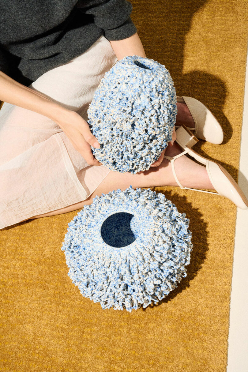 A person sitting on a tan carpet holds a textured, blue and white sculptural object; a similar object rests on the floor nearby.