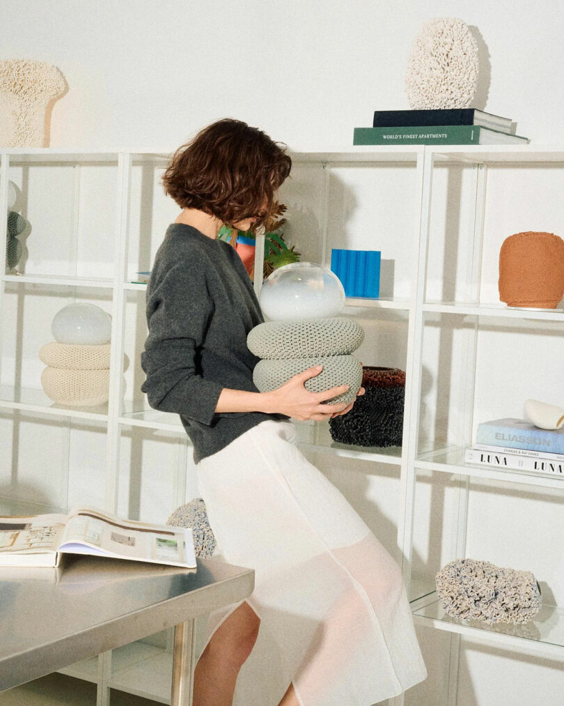 A woman in a grey sweater and white skirt stands by white shelves, holding a textured decorative object with a round glass top in a modern room.