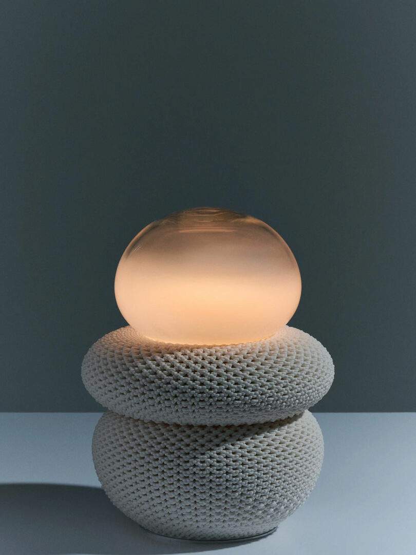 A modern lamp with a textured white base and a smooth, rounded, glowing glass top, set against a plain gray background.