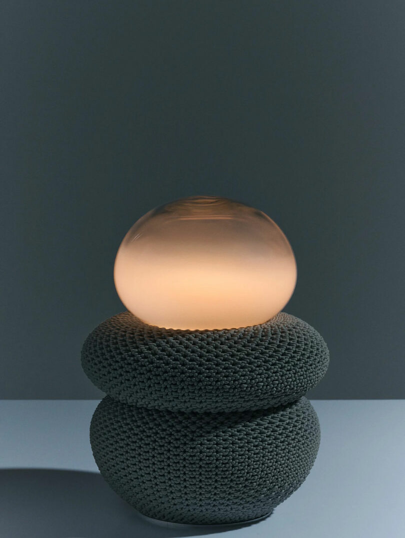 A round, softly glowing glass orb sits atop two stacked, textured gray spheres on a flat surface against a plain, dark background.