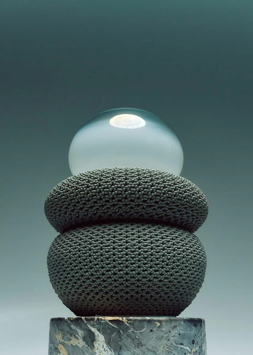 A spherical glass orb sits atop two stacked, textured, dark gray cushions on a marble base, against a gradient background.
