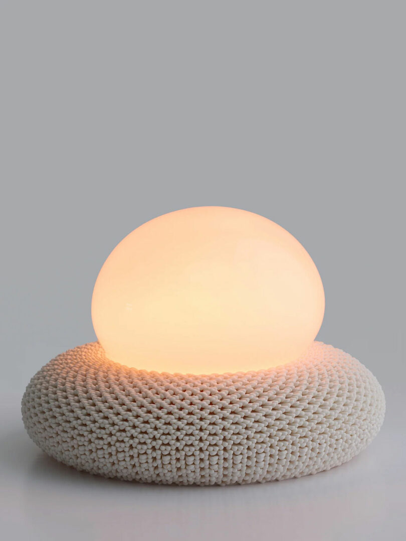 A round, glowing white lamp rests on a knitted, circular white base against a plain gray background.