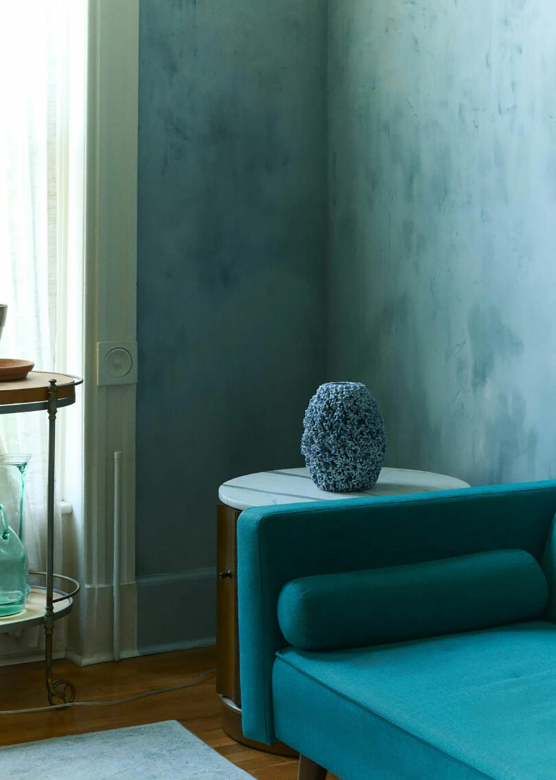 A blue sofa sits in front of a round white side table with a textured blue vase; light filters through a window with sheer curtains.