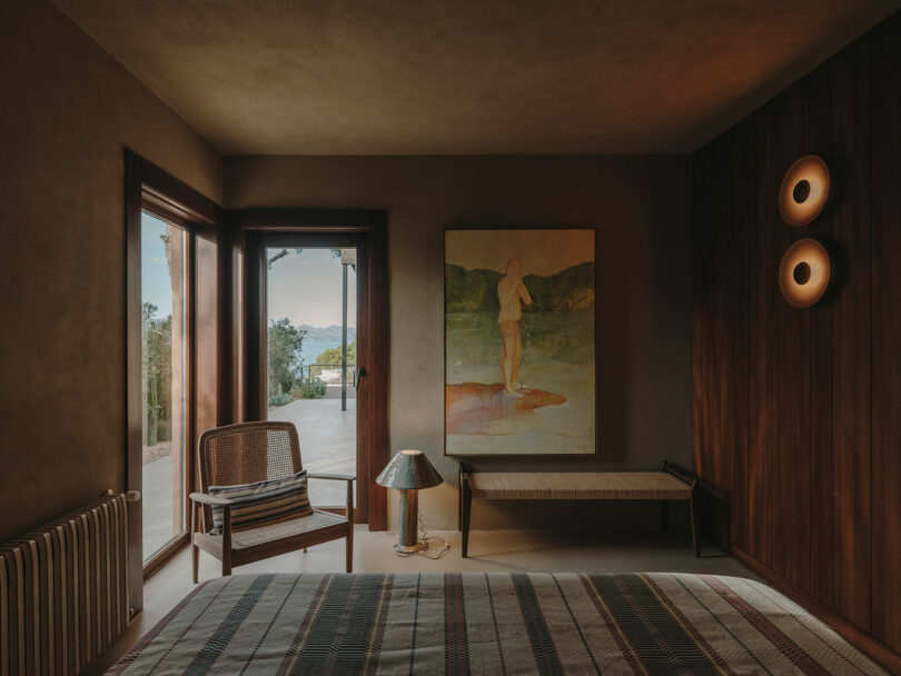 Un dormitorio con una iluminación cálida, una cama, una silla, un banco tejido, una lámpara, un gran cuadro en la pared y una puerta de cristal que conduce a una terraza exterior con un hermoso paisaje.