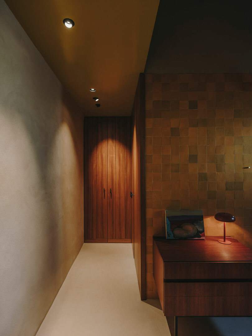 Un pasillo con paredes beige y focos en el techo conduce a una puerta de madera; Contra la pared de azulejos de la derecha hay una mesa de madera con una luz roja.