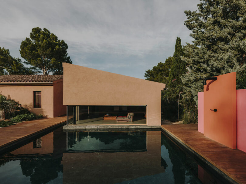 Una casa moderna con techo inclinado y grandes ventanales se encuentra junto a una piscina rectangular, rodeada de árboles y una terraza de madera bajo un cielo parcialmente nublado.