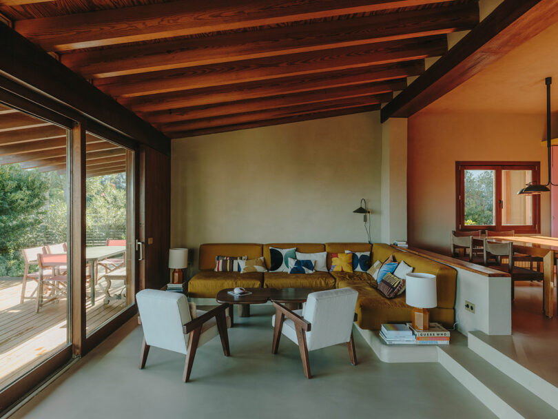 La moderna sala de estar cuenta con vigas de madera a la vista, un sofá seccional de color mostaza con almohadas, dos sillones blancos, una mesa de café redonda y grandes ventanales que se abren a una terraza con asientos al aire libre.