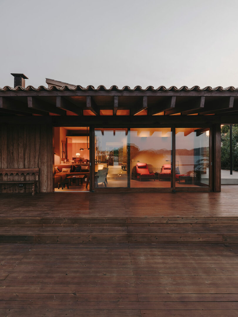 Vista exterior de una moderna casa de madera con grandes puertas y ventanas de cristal que muestran una cálida luz interior y reflejos exteriores al atardecer.