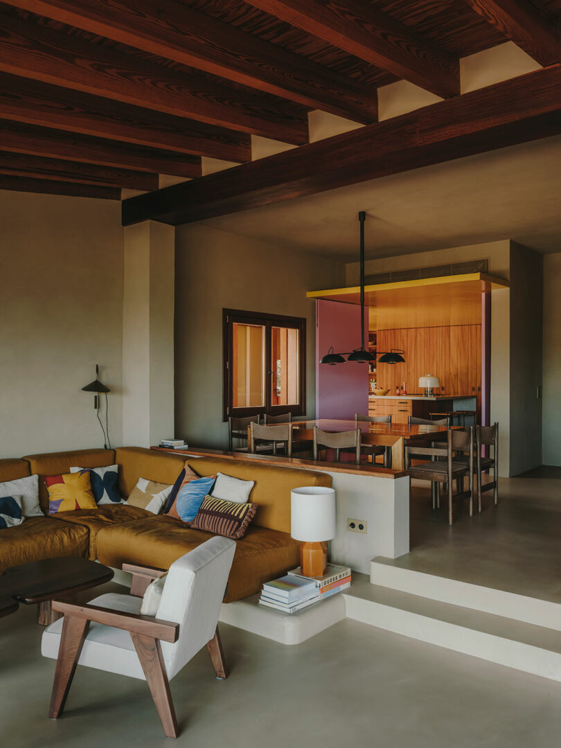 Moderna sala de estar y comedor con sofá seccional color mostaza, muebles de madera, techo con vigas a la vista y cocina con paredes coloridas al fondo.